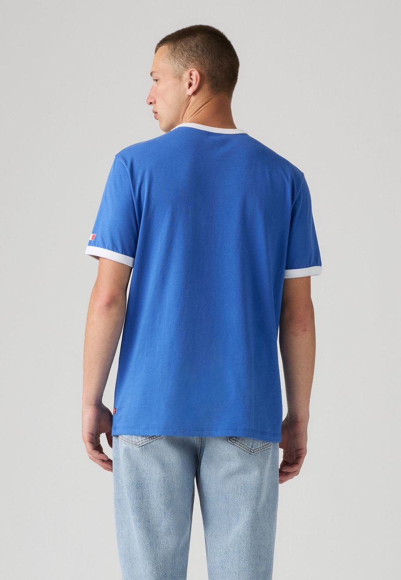 Uomo che indossa una camicia blu a maniche corte con bordi bianchi e jeans azzurri, in piedi di spalle su uno sfondo semplice.