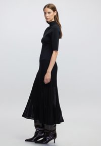 Haut noir à rayures associé à une jupe plissée fluide noire. Le mannequin porte des bottes noires à bout pointu et à brillance élevée. Design simple et épuré.