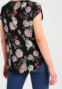 Blusa negra con flores y mangas cortas, que presenta un estampado de flores multicolores, un detalle de abertura en la espalda y un corte relajado. Combinada con vaqueros azules.