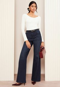Frau, die ein weißes Langarmoberteil, hoch taillierte dunkelblaue Jeans mit weitem Bein, schwarzen Gürtel, braune spitze Absätze trägt und eine rote Handtasche hält.