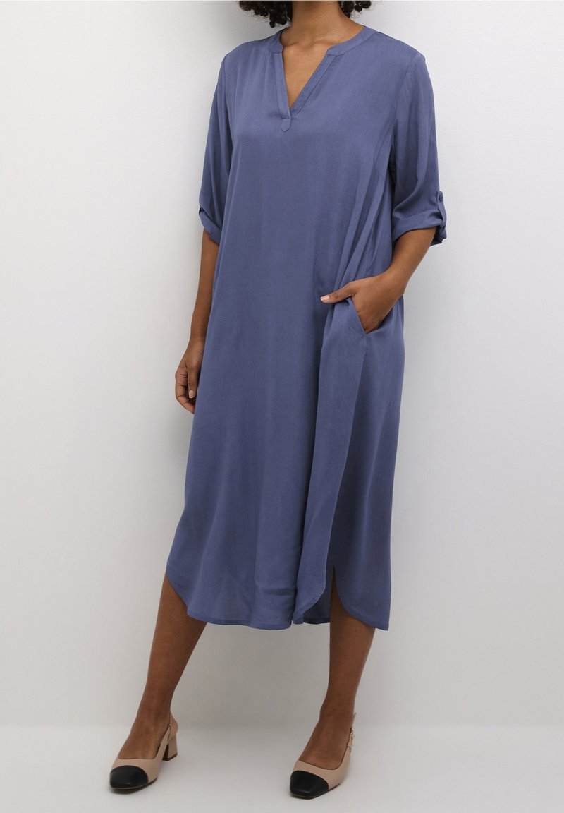 Femme portant une robe bleue ample arrivant au genou, manches retroussées, et des chaussures à talons carrés beiges avec des bouts noirs.