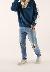 Felpa con zip blu e panna con colletto a quarti, abbinata a jeans chiari strappati e sneakers beige. Abbigliamento casual esposto.