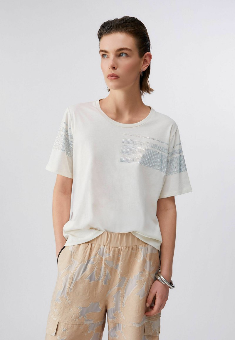 Jeune personne aux cheveux courts portant un t-shirt blanc avec des rayures pailletées et un pantalon beige à motifs floraux, debout les mains dans les poches.