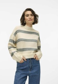 Maglione crema con strisce orizzontali grigie, collo alto e polsini a costine, abbinato a jeans blu a vita alta. Tessuto morbido, vestibilità rilassata.