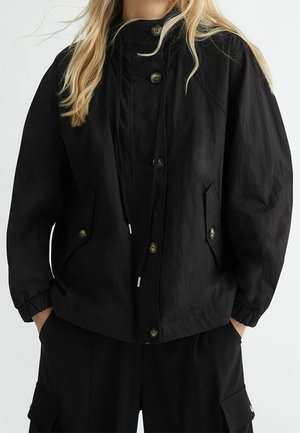 Person wearing a black buttoned jacket with elastic cuffs and drawstrings, hands in pockets, paired with black cargo pants.