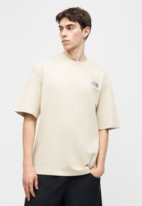 Νέος άντρας που φοράει ένα μπεζ oversized T-shirt με το λογότυπο The North Face, μαύρο σόρτς, κοιτάζοντας πλάγια μπροστά σε απλό φόντο.