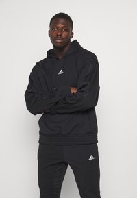 Zwarte Adidas hoodie met een trekkoord, ribbelmanchetten en -zoom; voorzien van het witte Adidas-logo. Gecombineerd met bijpassende zwarte broek.