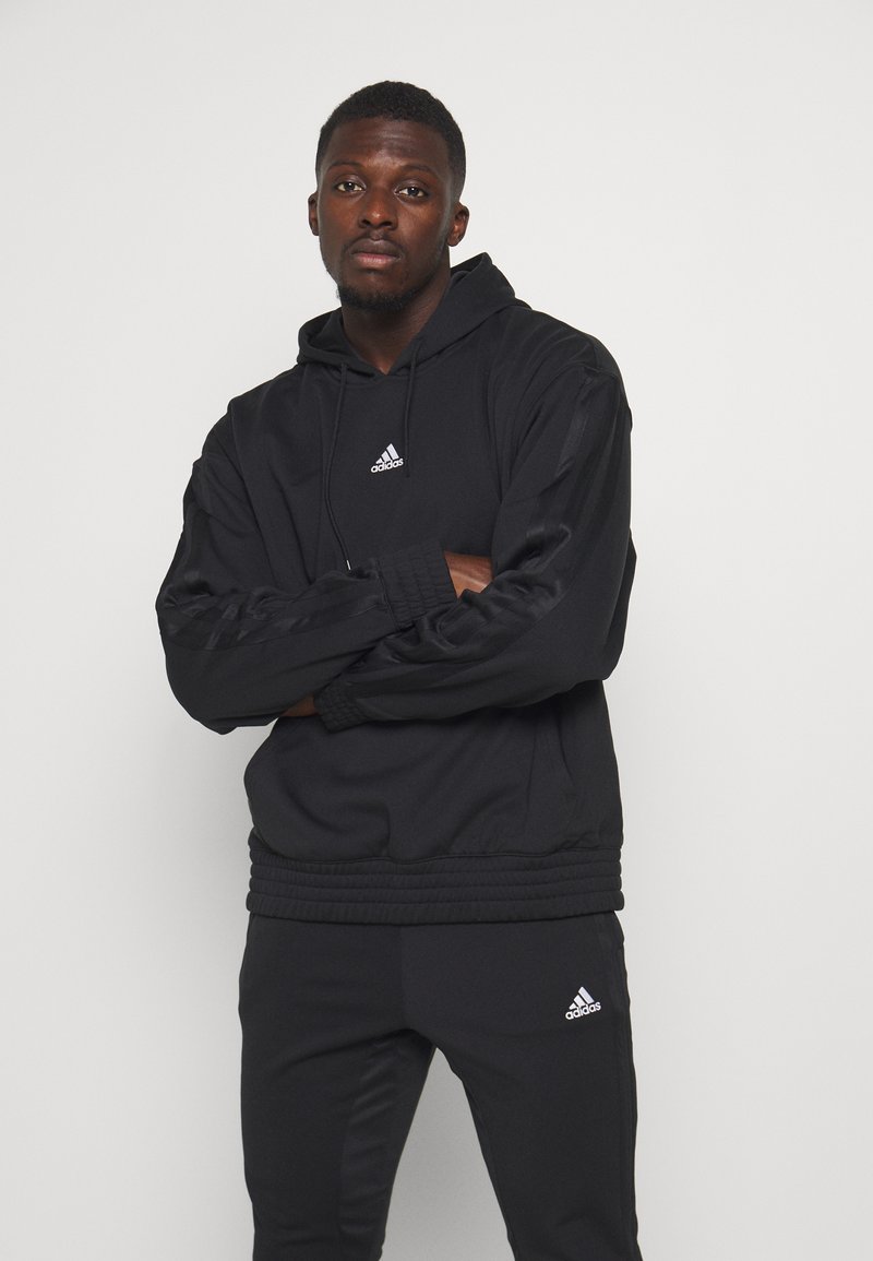 Zwarte Adidas hoodie met een trekkoord, ribbelmanchetten en -zoom; voorzien van het witte Adidas-logo. Gecombineerd met bijpassende zwarte broek.