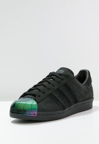 Sneaker in camoscio nero con tre strisce nere, caratterizzato da una punta lucida verde e viola. Superficie testurizzata e suola piatta nera.