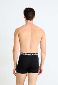 Schwarze Boxershorts mit einem breiten navyblauen Bund, auf dem "BJÖRN BORG" in weißer Schrift steht. Glatter Stoff mit enger Passform, von hinten betrachtet.