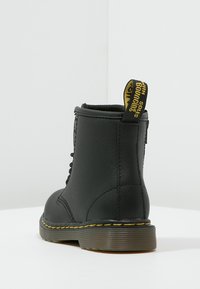Botină neagră din piele cu șireturi, cusături galbene și un tab de prindere cu branding "Dr. Martens" pe o talpă texturată.