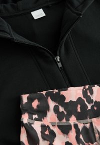 Veste à zip noire avec une texture lisse, associée à des leggings à imprimé léopard rose et noir, dotés d'une taille élastique et de coutures apparentes.