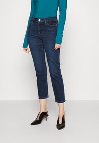 Jeans de mezclilla oscura con cintura alta y longitud de tobillo recortada. Llevados con una blusa de manga larga en color verde azulado y tacones negros de punta.