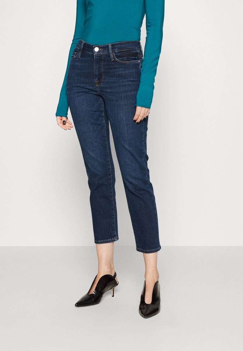 Jeans de mezclilla oscura con cintura alta y longitud de tobillo recortada. Llevados con una blusa de manga larga en color verde azulado y tacones negros de punta.