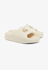 Beige slip-on slides made of rubber with a textured footbed, featuring an open toe and a crocodile logo in tan on the strap.