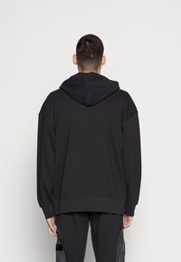 Man som står vänd mot vit vägg, iförd svart oversize-hoodie med huven nere och svarta byxor med sidofickor.