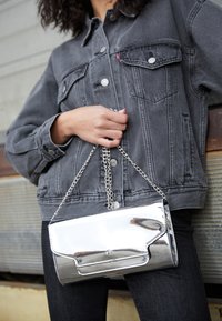 Silverfärgad metallisk clutchväska med rektangulär form, försedd med en kedjesnodd och ett klafflock. Bärs av en person i en grå denimjacka.