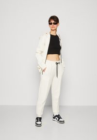 Sweat à capuche zippé blanc avec un crop top noir, un jogging blanc assorti avec un cordon de serrage noir, et des baskets noir et blanc. Textures lisses.