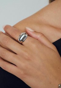 Silverring med en slät, rundad design på ett finger, med en glansig finish och subtil textur, bärs på en bar hand.