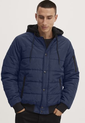 Giacca a vento trapuntata blu con dettagli neri, cappuccio, tasche con zip e bottoni a pressione. La texture leggermente imbottita migliora il calore.