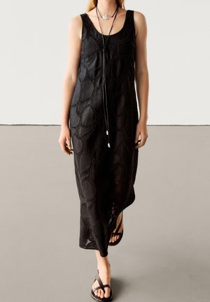 Femme portant une robe midi noire texturée sans manches, des sandales noires à brides et un long collier noir avec un pendentif argenté.