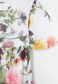 Das blumenbedruckte Kleid hat einen weißen Grundton mit verschiedenen Blumen in Rosa, Gelb und Lila, sowie gepufften Ärmeln und einem gerafften Saum.