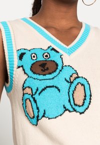 En bomullsstickad väst med en tecknad björngrafik i blått och brunt på en krämfärgad bakgrund, med turkosa ribbade detaljer runt halsringningen och ärmhålen.