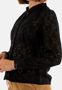 Blusa nera in pizzo con colletto scallop e maniche lunghe, dotata di chiusura frontale a bottoni e dettagli florali in trasparenza.