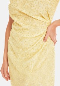 Gelbes Kleid aus strukturiertem Stoff mit zarter floralem Stickerei und drapierter Gestaltung, mit einer Hand in einer Seitentasche.
