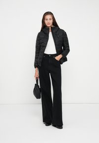 Black quilted puffer jacket with a high collar, paired with a white top and black wide-leg jeans. Model holds a small black bag.