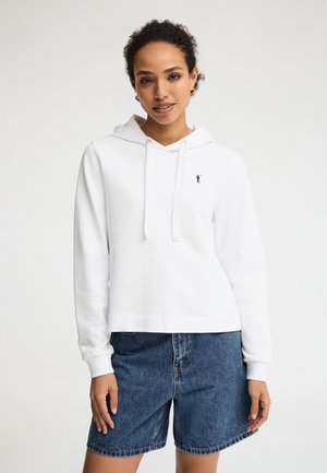 Kvinde iført en hvid hættetrøje med snore og et lille logo på brystet, kombineret med blå denimshorts, står mod en hvid baggrund.