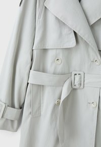 Trench-coat gris clair avec larges revers, boutons croisés, taille ceinturée et détail de sangle sur la manche.