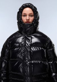 Musta pehmeä puffer-takki kiiltävällä pinnalla, korkealla kauluksella ja huppulla. Sisältää suuret tikatut osat ja suljetun vetoketjun.