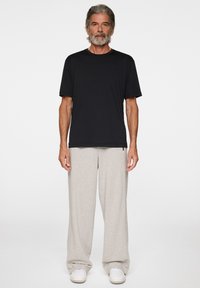 Man med grått hår och skägg som bär en svart kortärmad skjorta, ljusgrå vida byxor och vita sneakers, stående mot en enkel bakgrund.