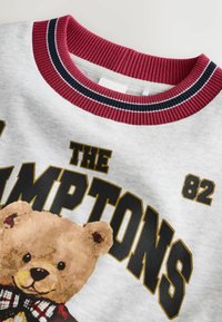 Szary sweatshirt z okrągłym bordowym ściągaczem, z grafiką przedstawiającą misia pluszowego oraz wyraźnym czarnym napisem "THE HAMPTONS 82".