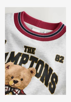 Sivkasta srajca s okroglo bordo rebrasto ovratnikom, ki vsebuje grafiko medvedka in bold črn napis "THE HAMPTONS 82".