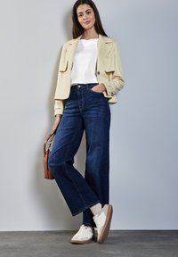 Beige Jacke, weißes T-Shirt, dunkelblaue weit geschnittene Jeans und weiße Sneakers mit braunen Akzenten. Enthält eine braune Handtasche und eine glatte Textur.