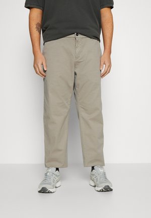 Personne portant un pantalon gris clair, des chaussettes noires, des chaussures de sport grises et une chemise foncée, debout devant un fond blanc uni.