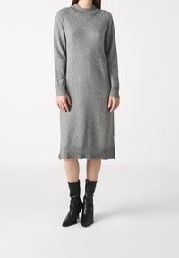 Abito grigio a maglia con colletto alto, maniche lunghe e orlo a coste. Indossato con stivali neri aderenti alla caviglia. Tessuto morbido e silhouette rilassata.
