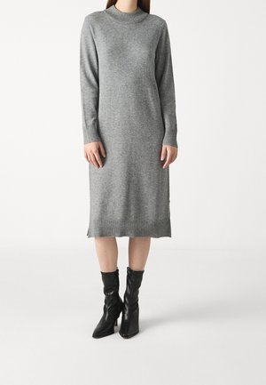 Abito grigio a maglia con colletto alto, maniche lunghe e orlo a coste. Indossato con stivali neri aderenti alla caviglia. Tessuto morbido e silhouette rilassata.