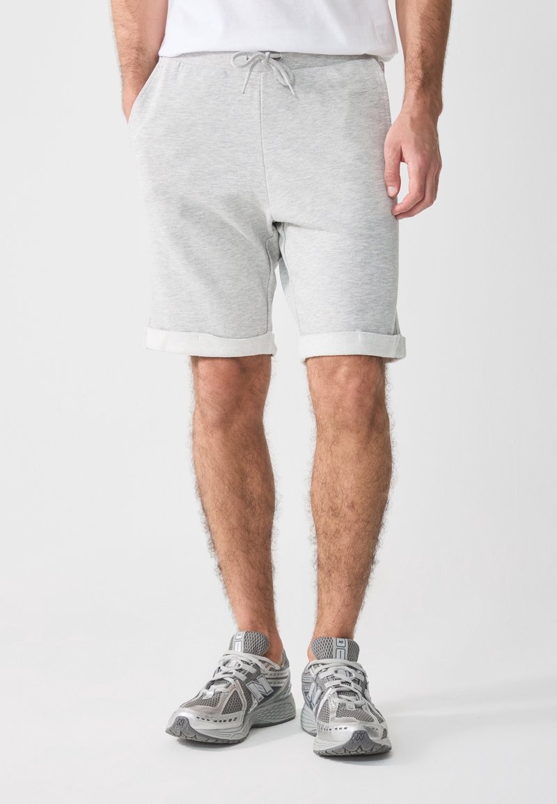 Homme portant un short gris clair avec cordon de serrage et revers roulés, un t-shirt blanc et des chaussures de course grises, debout devant un fond uni.