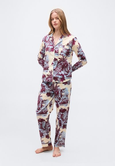 Νεαρή γυναίκα στέκεται ξυπόλυτη φορώντας ταίριασμα κρεμ πιτζάμες με μαρόν και μπλε λουλουδένιο και ζωικό μοτίβο, με τα χέρια πίσω από την πλάτη, ουδέτερη έκφραση.