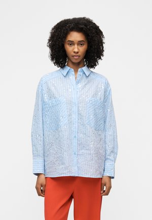 Femme aux cheveux bouclés portant une chemise boutonnée transparente bleu clair ornée de sequins et de grandes poches, associée à un pantalon orange vif.
