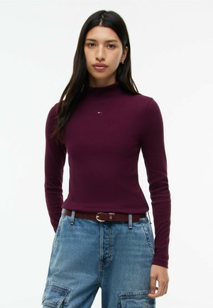 Donna con i capelli lunghi e scuri indossa un maglione aderente a collo alto color bordeaux e pantaloni cargo in denim celeste con cintura marrone.