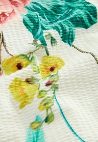 Tissu à motifs floraux présentant des fleurs jaunes avec des centres marron, des feuilles vertes et des accents turquoise sur un fond blanc texturé.