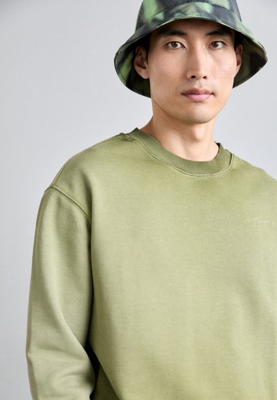 Felpa in cotone verde oliva con colletto a girocollo e polsini a costine, con un delicato ricamo. Abbinata a un cappello a secchiello con motivo.