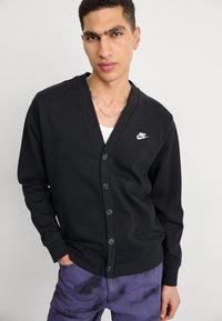 Svart kofta med V-ringning, knappstängning, långa ärmar och en liten vit Nike-logotyp på vänster bröst. Slät textur, avslappnad design.