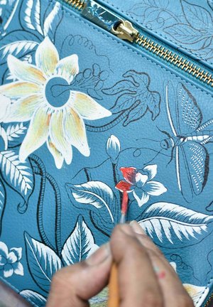 Blauwe leren oppervlakte met bloemmotief. Handgeschilderde details van rode bloemen met een fijne kwast. Gouden rits zichtbaar aan de bovenkant.