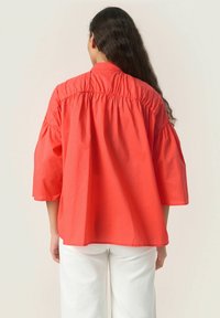 Blusa in cotone corallo con schiena arricciata, maniche a sbuffo corte e orlo dritto, abbinata a pantaloni bianchi. La superficie appare liscia.