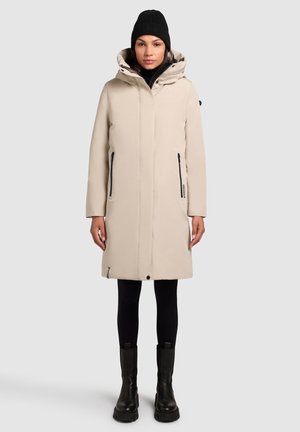 Parka imperméable beige avec capuche, dotée de poches latérales zippées, d'une coupe droite et d'une texture douce, associée à des leggings noirs et des bottes.